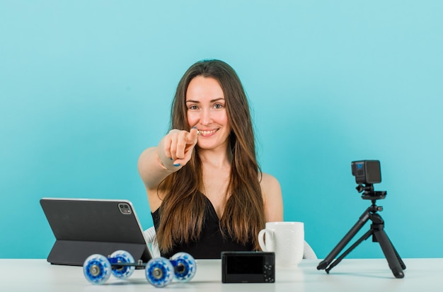 Smiling blogger girl is looking at camera by pointing camera with forefinger on blue background