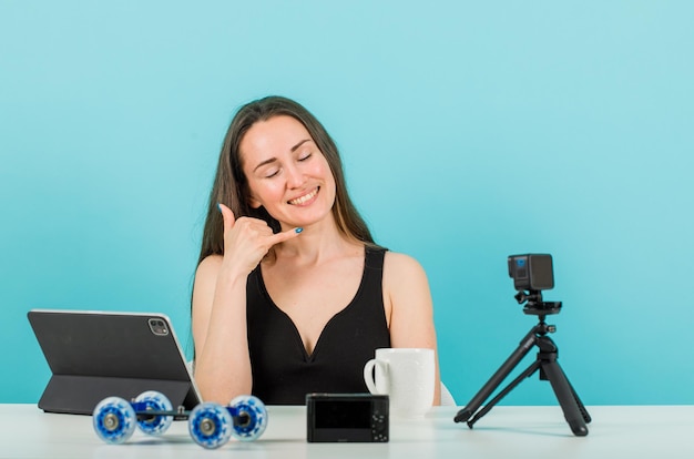 Smiling blogger girl is posing at camera by showing mobile gesture with hand on blue background