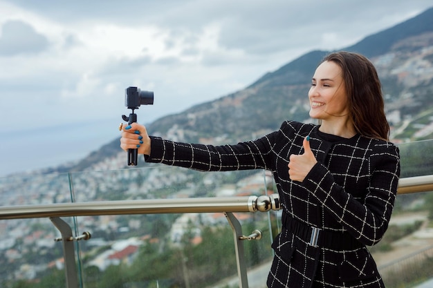 Smiling blogger girl is taking selfie with her mini camera by showing perfect gesture against the background of city view