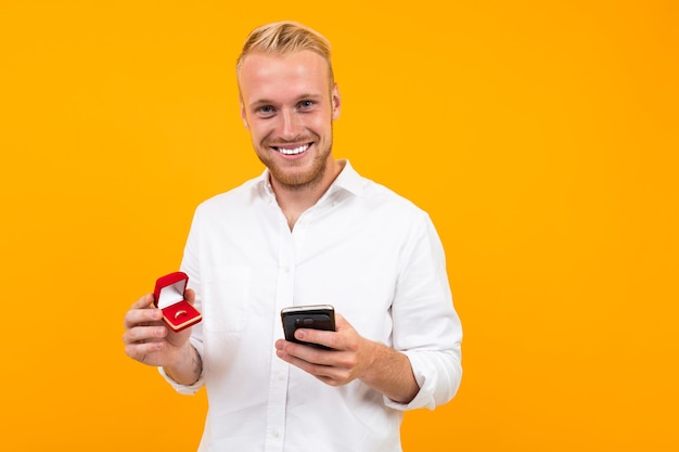 Smiling blond european man makes an offer holding a ring in a box and a phone on a yellow background.