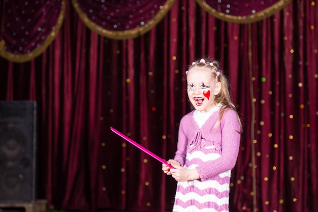 Smiling Blond Girl Wearing Clown Make Up and Striped Dress Standing on Stage Holding Over Sized Pink Comb in front of Red Curtain