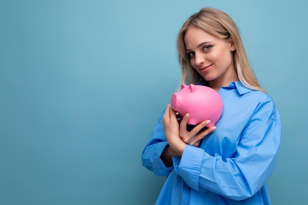 Smiling blond young adult hugging a piggy bank where she keeps savings on a blue isolated background