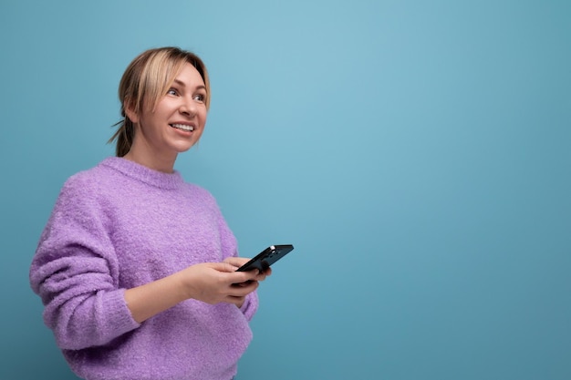 Smiling blond young woman in a purple hoodie with a smartphone in her hands on a blue background