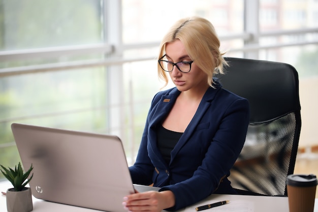 Smiling blonde businesswoman with glasses using laptop in the office.