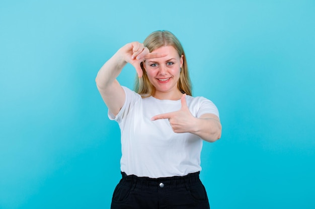 Smiling blonde girl is hsowing frame gesture on blue background