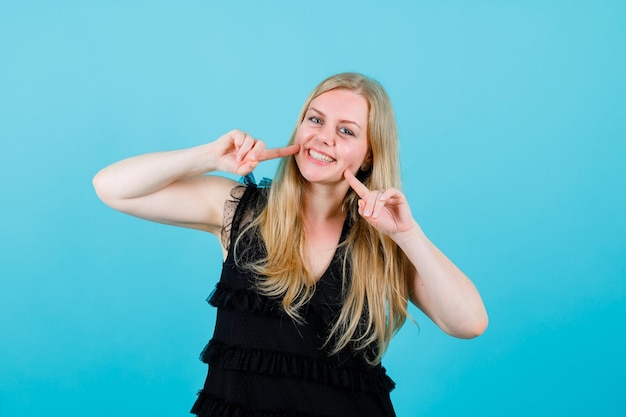 Smiling blonde girl is looking at camera by holding forefingers on cheeks on blue background