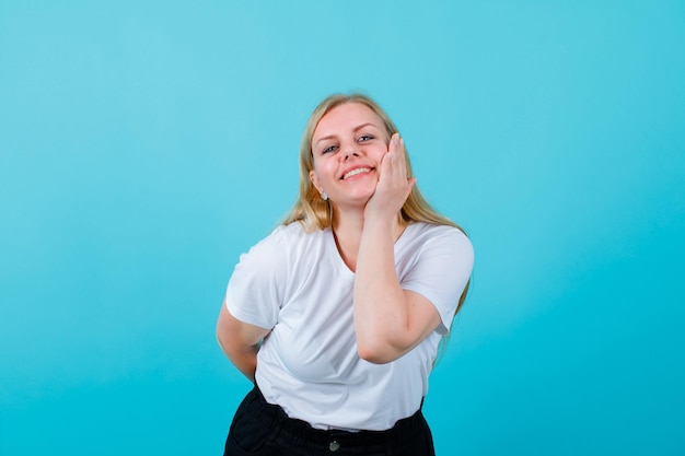 Smiling blonde girl is posing to camera by putting hand on cheek on blue background