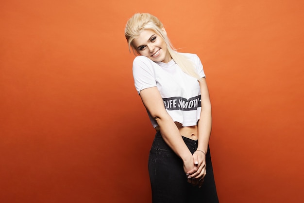 Photo smiling blonde model with perfect body in black jeans and white t-shirt posing at orange wall,