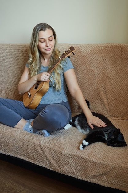 Smiling blonde petting her cat while sitting on the couch at home holding a ukulele in her hands