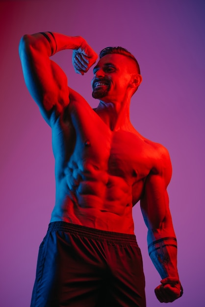 A smiling bodybuilder with a beard is posing with a flexed bicep under the blue and red light