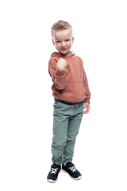 Smiling boy is standing A cute blond man with a trendy hairstyle in a brown hoodie and gray trousers Joy and positivity Full height Isolated on white background Vertical