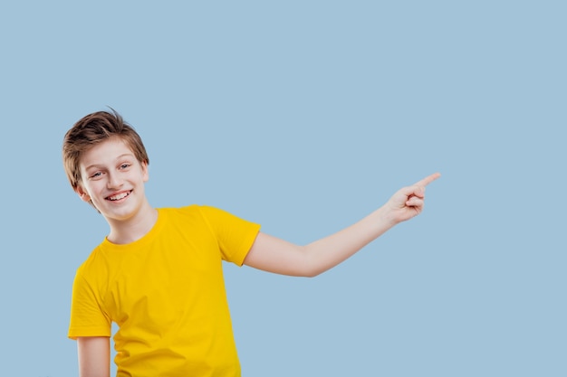 Smiling boy pointing his finger to the left side, in yellow t-shirt isolated on blue