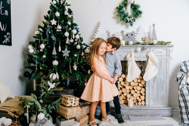 Smiling boy whispering to ear of his cute and beuatiful girlfriend in fron of cristmas tree with fireplace.