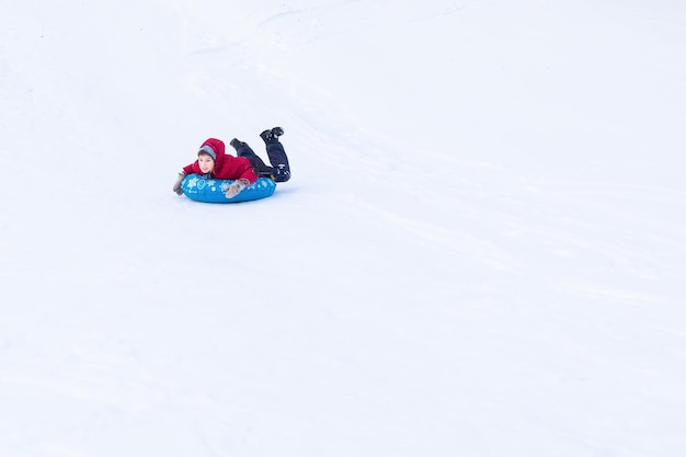 Smiling boy in winter clothes sliding on blue tubing in distance among white snow