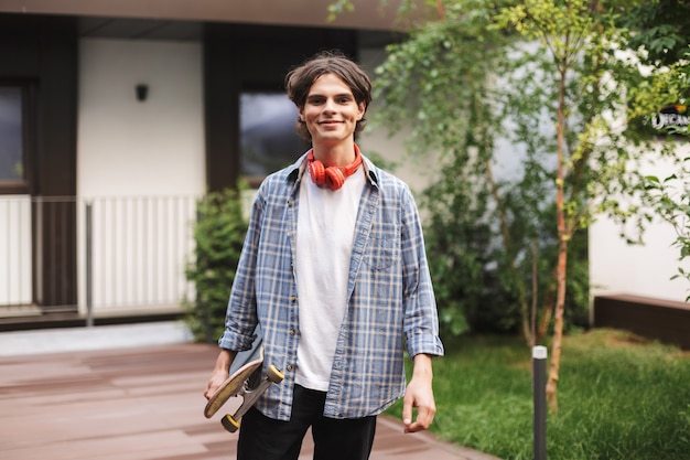 Smiling boy with red headphones standing and holding skateboard while joyfully 