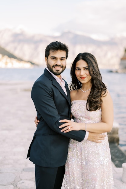 Smiling bride and groom stand hugging each other around the waist on a pier by the sea