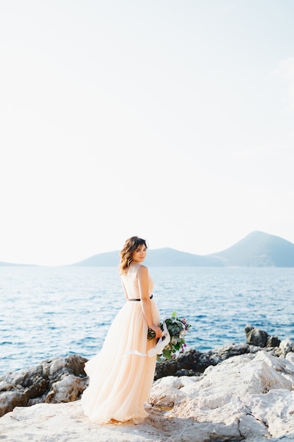 Smiling bride in a pastel wedding dress stands on a rock above the sea with a bouquet of flowers