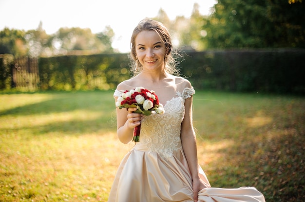 Smiling bride in a white dress holding a beauriful red roses bouquet in the green park. Conception of the wedding