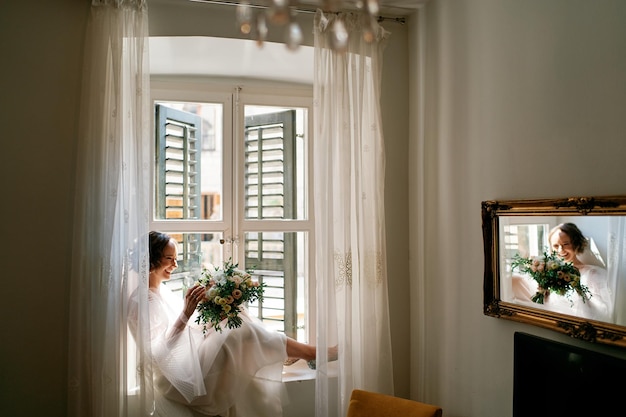 Smiling bride with a bouquet sits on the windowsill near the window reflected in the mirror on the