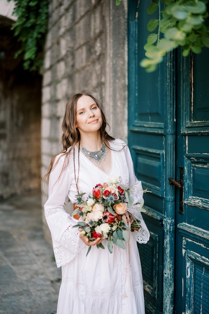 Smiling bride with a bouquet stands near the green wooden door of an old stone house