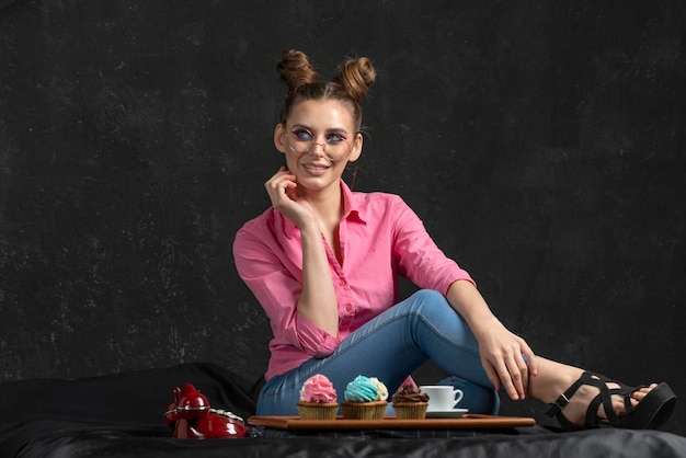 Smiling bright young woman in glasses and pink shirt on black background Tray of muffins with multicolored cream