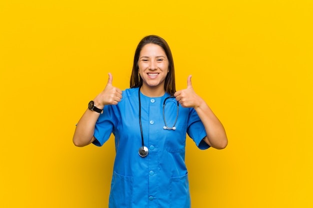 Smiling broadly looking happy, positive, confident and successful, with both thumbs up isolated against yellow wall