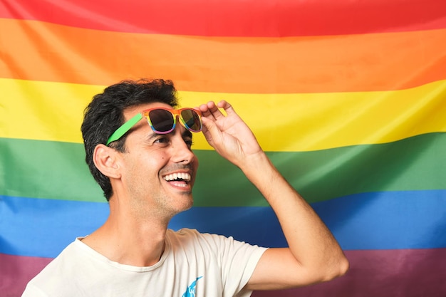 Smiling brunette boy in white tshirt with rainbow lgbtq glasses raising his glasses on gay pride rainbow flag background