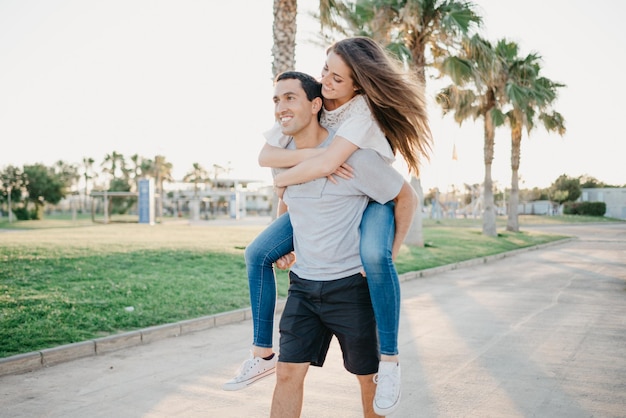 A smiling brunette girl is riding her Hispanic boyfriend between palms in Spain