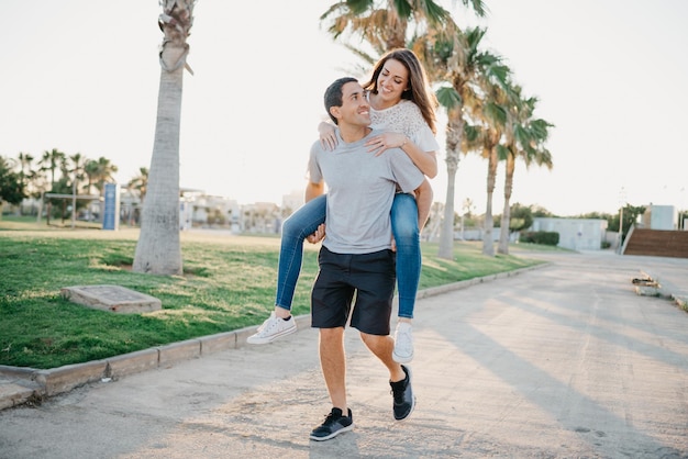 A smiling brunette girl is riding her Hispanic boyfriend between palms in Spain