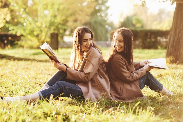 Smiling brunette twin girls sitting back to back on the grass and looking at each other, legs slightly bent in knees, with brown books in hands, wearing casual coat in autumn park on blurry background