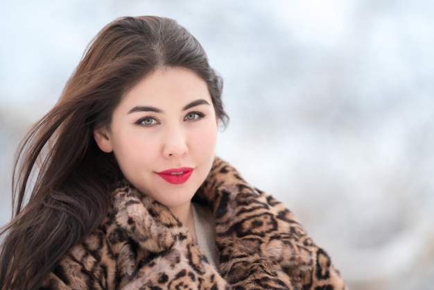 Smiling brunette with long hair dressed in faux fur coat with leopard pattern and looking at camera