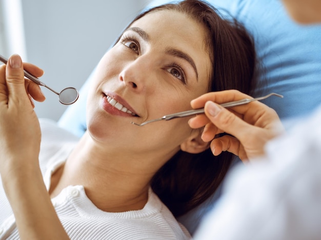 Smiling brunette woman being examined by dentist at dental clinic. Hands of a doctor holding dental instruments near patient's mouth. Healthy teeth and medicine concept.