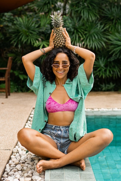 Smiling brunette woman  posing with pineapple sitting near swimming pool in tropical resort