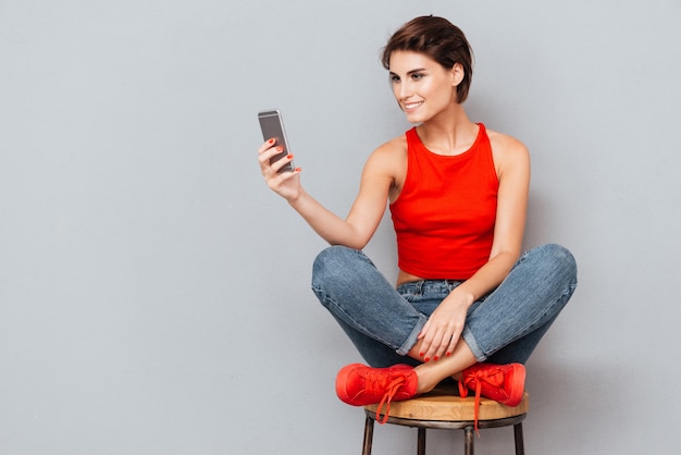 Smiling brunette woman sitting on the chair and using smartphone over gray background