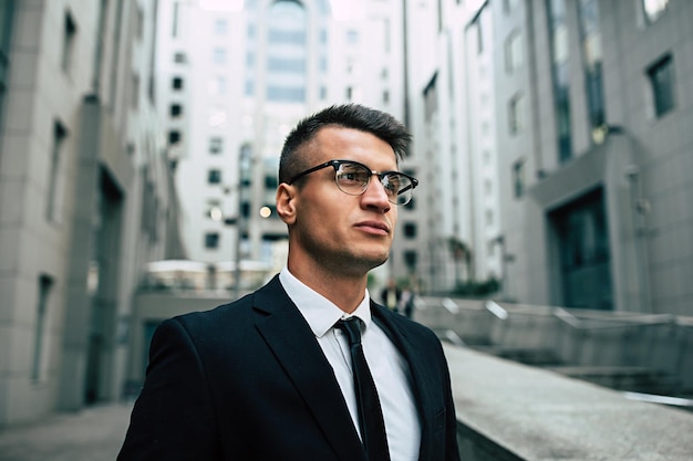 Smiling business man in full black suit and glasses looking on camera standing on cityscape background