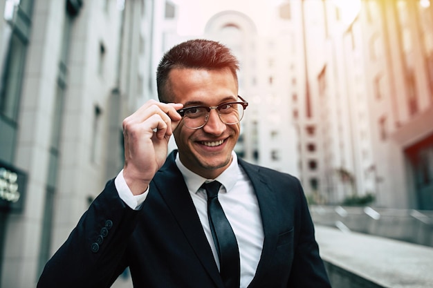 Smiling business man in full black suit and glasses looking on camera standing on cityscape background