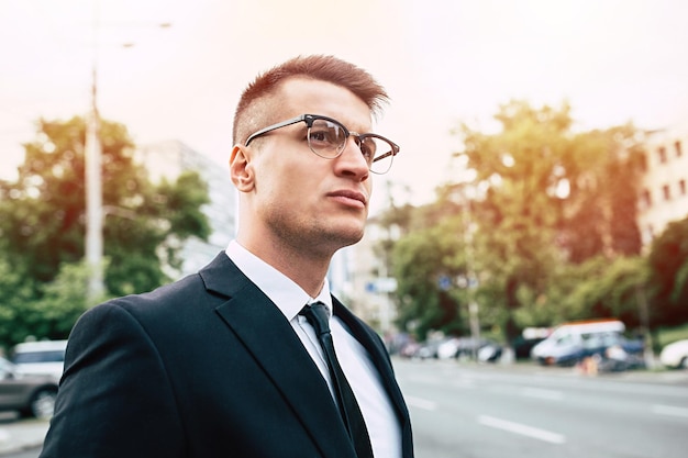 Smiling business man in full black suit and glasses looking on camera standing on cityscape background