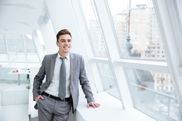 Smiling Business man standing near the window in office with arm in pocket and looking away