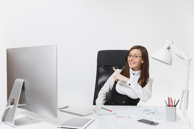 Smiling business woman sitting at the desk, working at computer with documents in light office