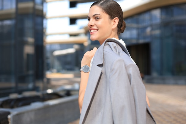 Smiling business woman standing with a jacket over her shoulder near office building.