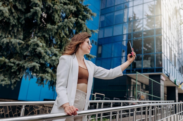 Smiling business woman in white suit and sunglasses making video call to friend using phone during break standing near modern office building