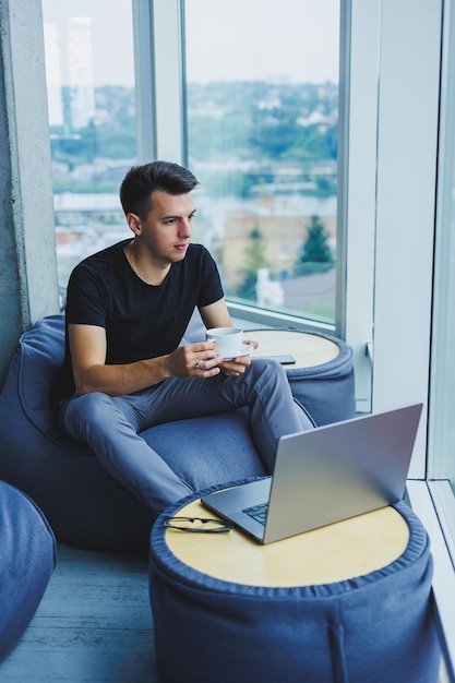 Smiling businessman looking for something and typing on a laptop and drinking fresh black coffee The concept of a modern successful person Young focused guy with glasses in an open office