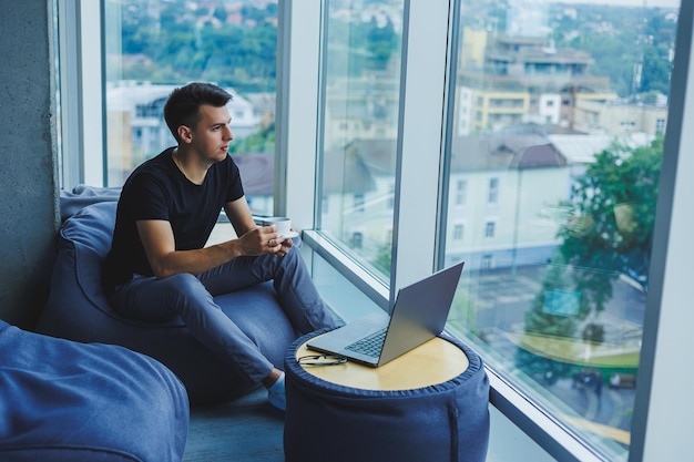 Smiling businessman looking for something and typing on a laptop and drinking fresh black coffee The concept of a modern successful person Young focused guy with glasses in an open office