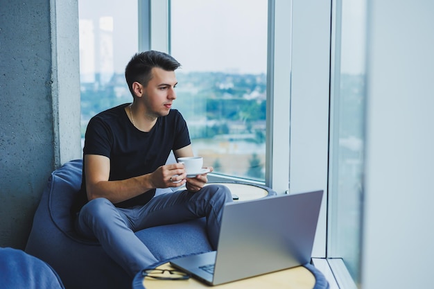 Smiling businessman looking for something and typing on a laptop and drinking fresh black coffee The concept of a modern successful person Young focused guy with glasses in an open office