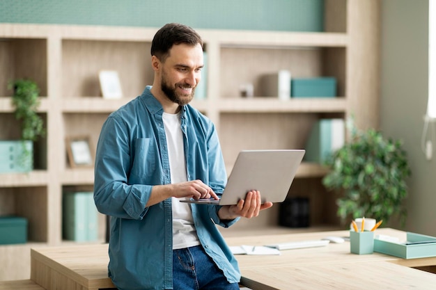 Smiling businessman using laptop computer while standing near desk in modern office