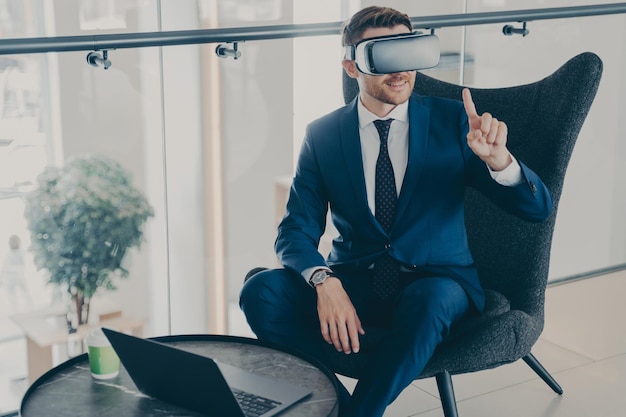 Smiling businessman using VR headset glasses while sitting in office lobby and working on laptop