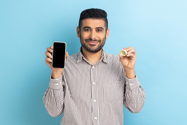 Smiling businessman with beard holding btc golden coin and cellphone looks at camera