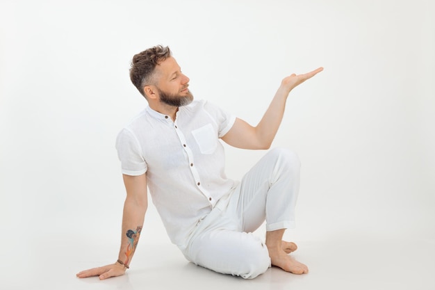 Smiling businessman with tattoo sit on the floor in white outfit on white background advertising man