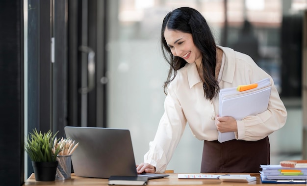 Smiling businesswoman holding stack of document and checking data on laptop computer standing at her desk office