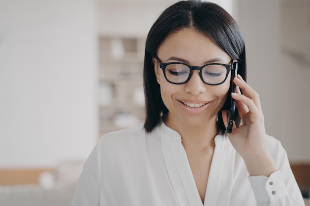 Smiling businesswoman wearing glasses talking on smartphone with client answering call in office